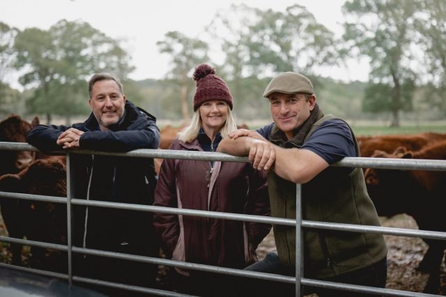 Pictured left to right – Richard Snelling, Sarah and Andrew Parry-Norton. They are standing behind a metal gate leaning over the top. An open field with cows is directly behind them