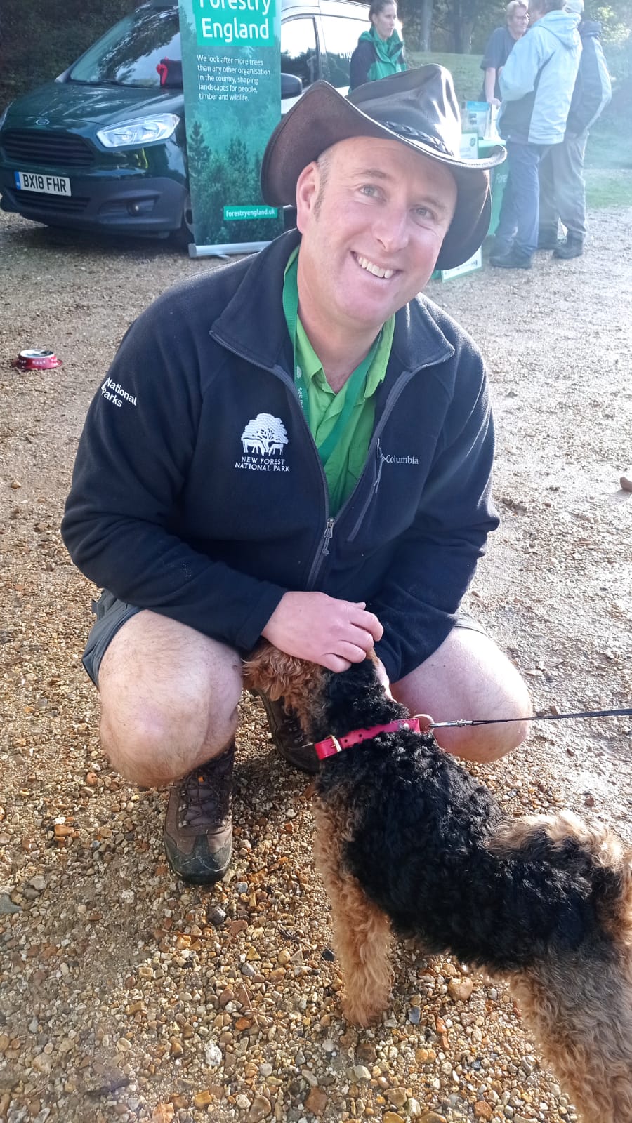 A New Forest National Park Authority staff member. He is wearing a hat, smiling and crouched down stroking a small dog. 