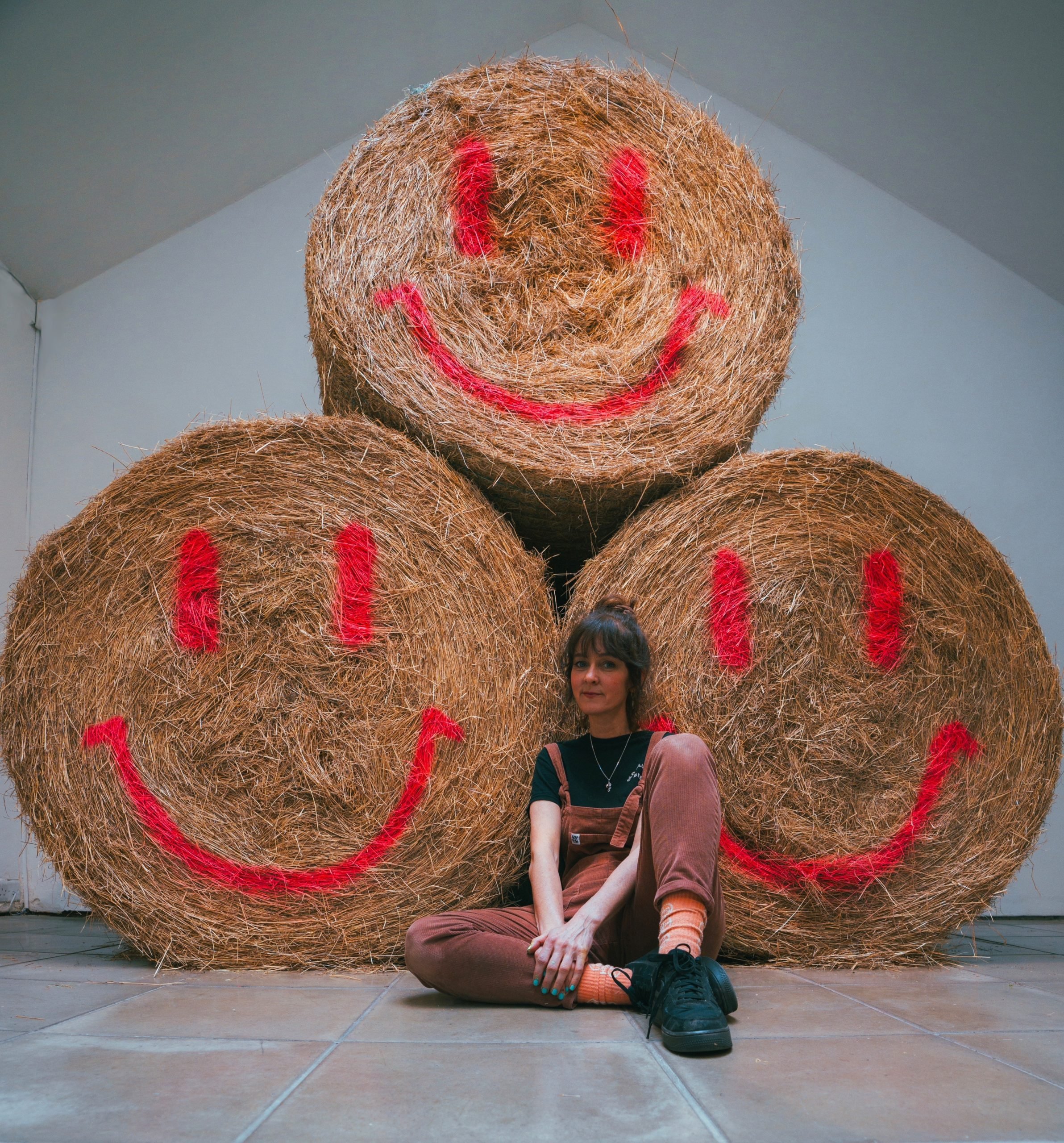 New Forest Artist in Residence Beccy Mccray sat in front of three hay bales with red spray painted smiley faces on the front. One hay bale is stacked on top of the other two.