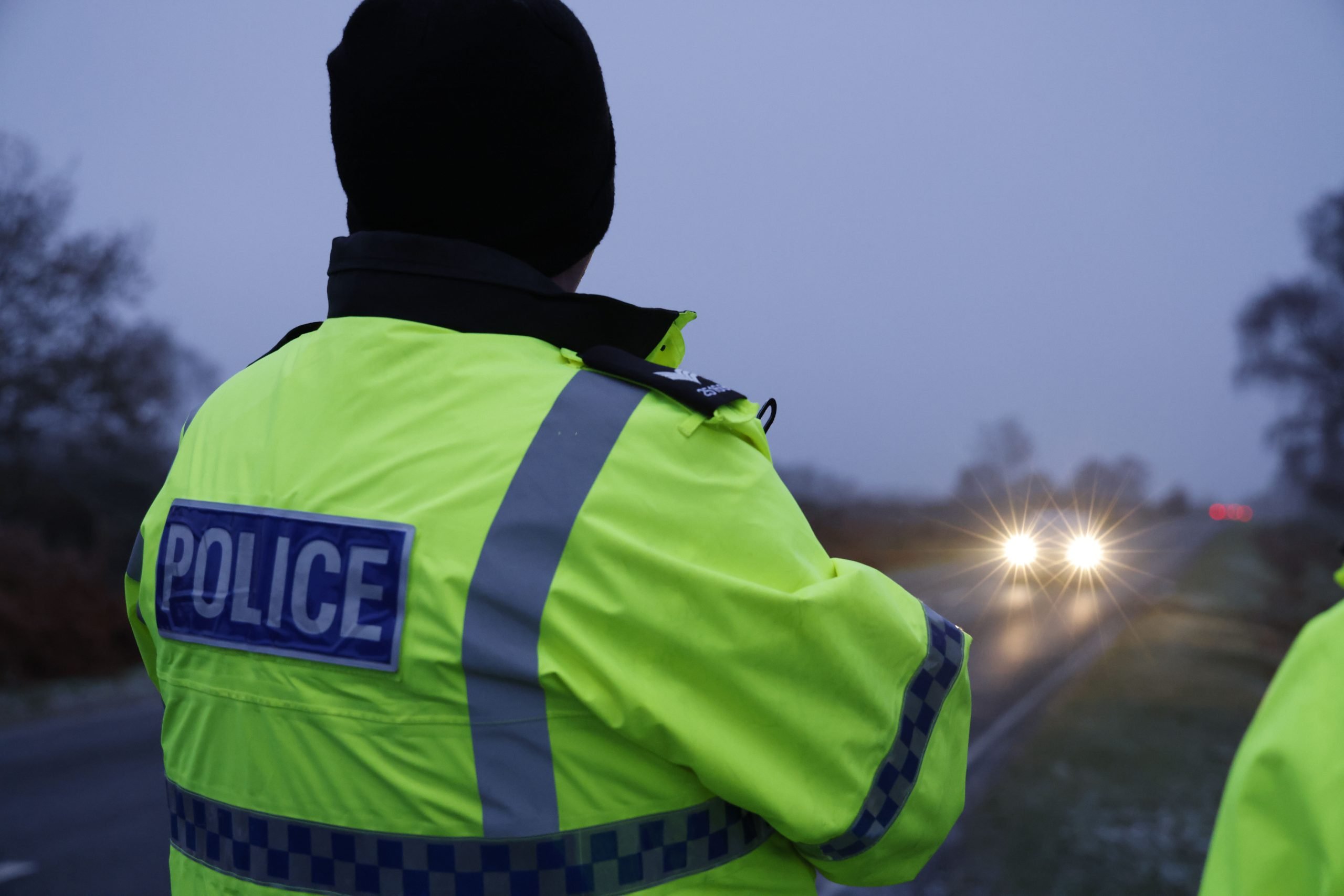 A policeman in bright fluorescent jacket with POLICE on the back. Stood on a roadside with arms folded and facing away from the camera. A car is oncoming and can be seen over his shoulder. It is early morning and the car has it's headlights on.