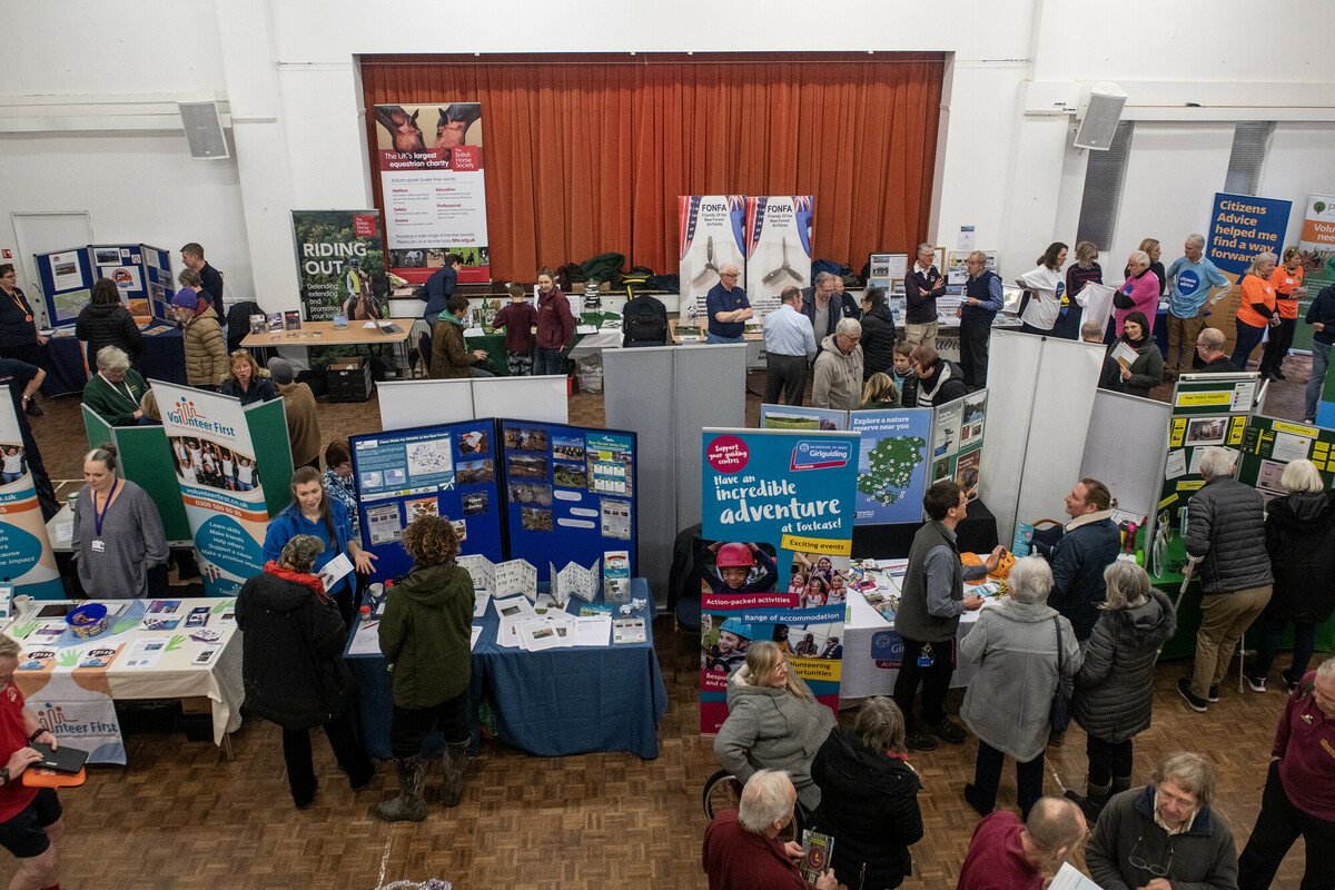 A busy village hall during the New Forest National Park Volunteer Fair 2023. Photo taken from high up looking down at three rows of stall holders and lots of people talking to the exhibitors and one another. Some are holding leaflets and handouts from the various volunteer opportunities.