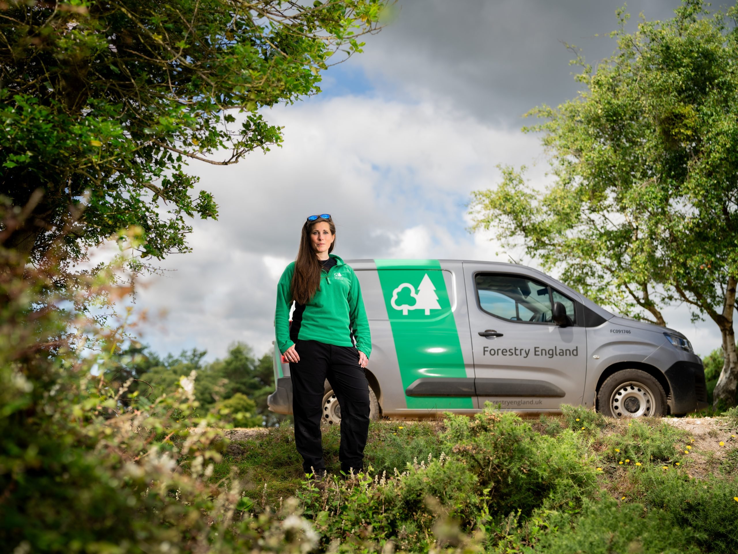 Forestry England ranger Charlotte Belcher stood in the open Forest in uniform, her ranger van is parked behind her on a track