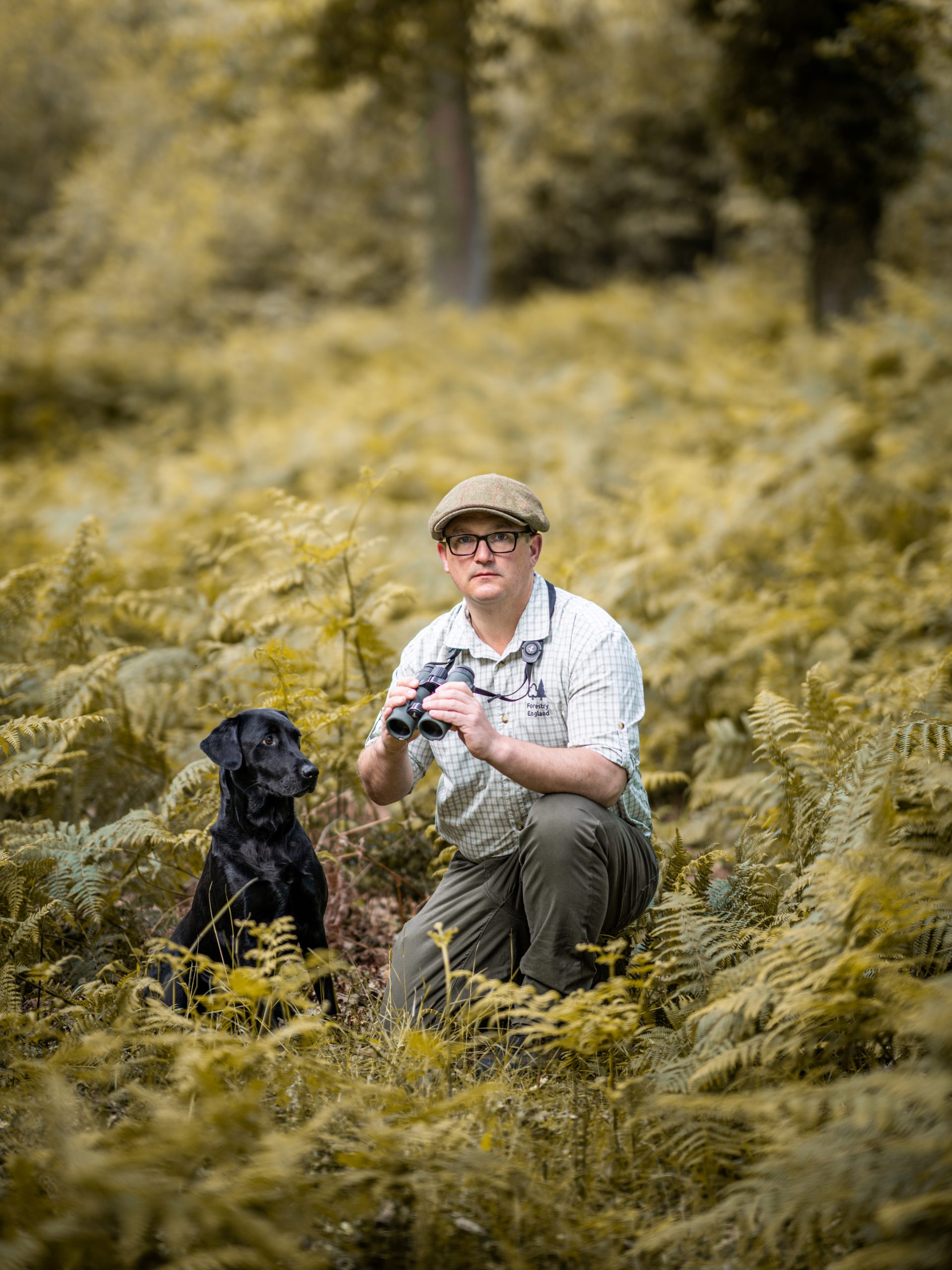 Forest Keeper Lee Knight crouched in woodland with his dog beside him, he is holding binoculars and looking off into the distance