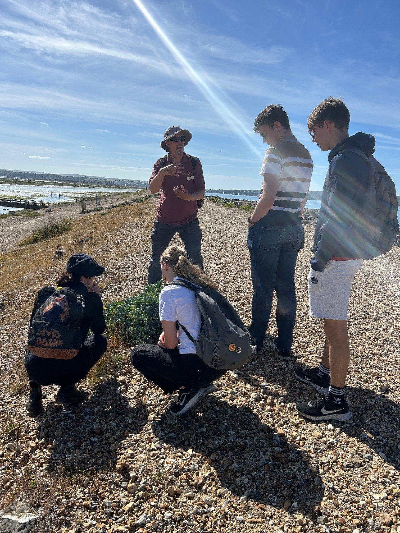 Coastal erosion session with CET, Hurst Spit (credit TFL) A group of young people learning about coastal erosion at Hurst Spit, some are standing, some are crouching on the pebbles, it's a sunny day
