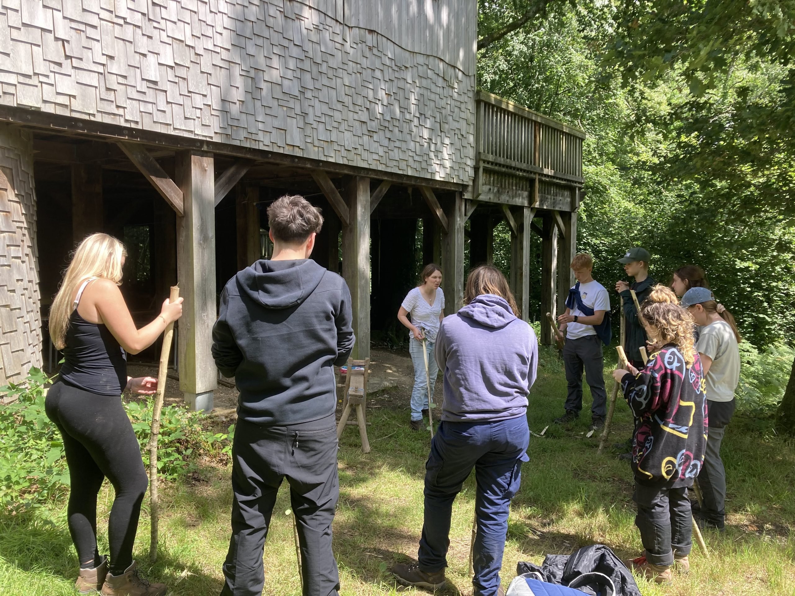 Listening to input on how to use shave horse and draw knives for carving shepherding sticks (credit CET) Young people stood with shepherding sticks listening to an adult giving a carving tutorial, they are all outside