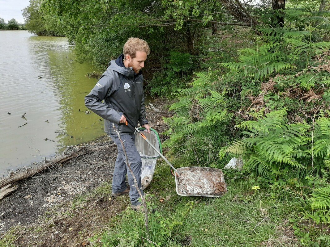 Former apprentice ranger Jake lifts a used displosable bbq into a plastic bag with a litter picker. He is stood by a river side