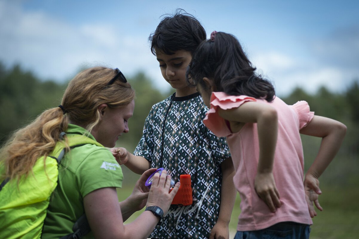 NPA ranger Hayden with two young children looking at a bug through a magnifying pot on a guided walk for Southampton and Winchester Visitors Group who support people with refugee status.