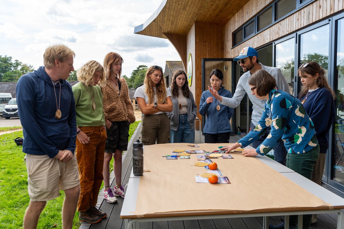 Jim from Decarbon Easer leads an interactive climate fresk session with the New Forest Youth Board Jim from Decarbon Easer leads an interactive climate fresk session with the New Forest Youth Board. The members are stood around a table outside the front of the Fort Climate Centre building, there are cards depicting aspects of climate change on the table.