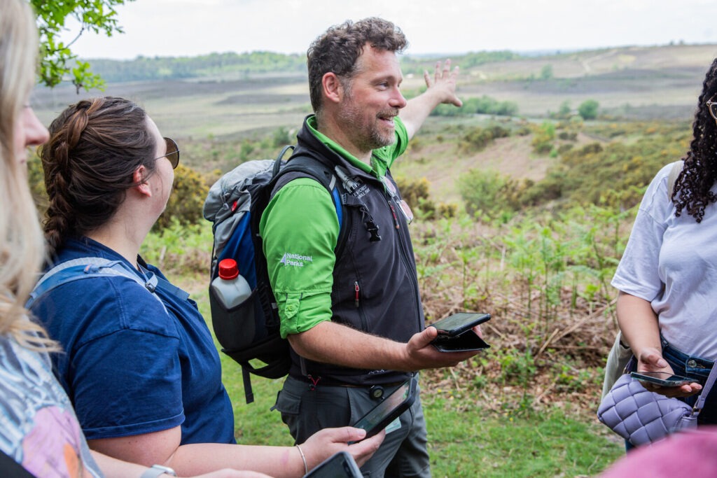 A man holding a phone and wearing a back pack pointing to a hillside with three people nearby
