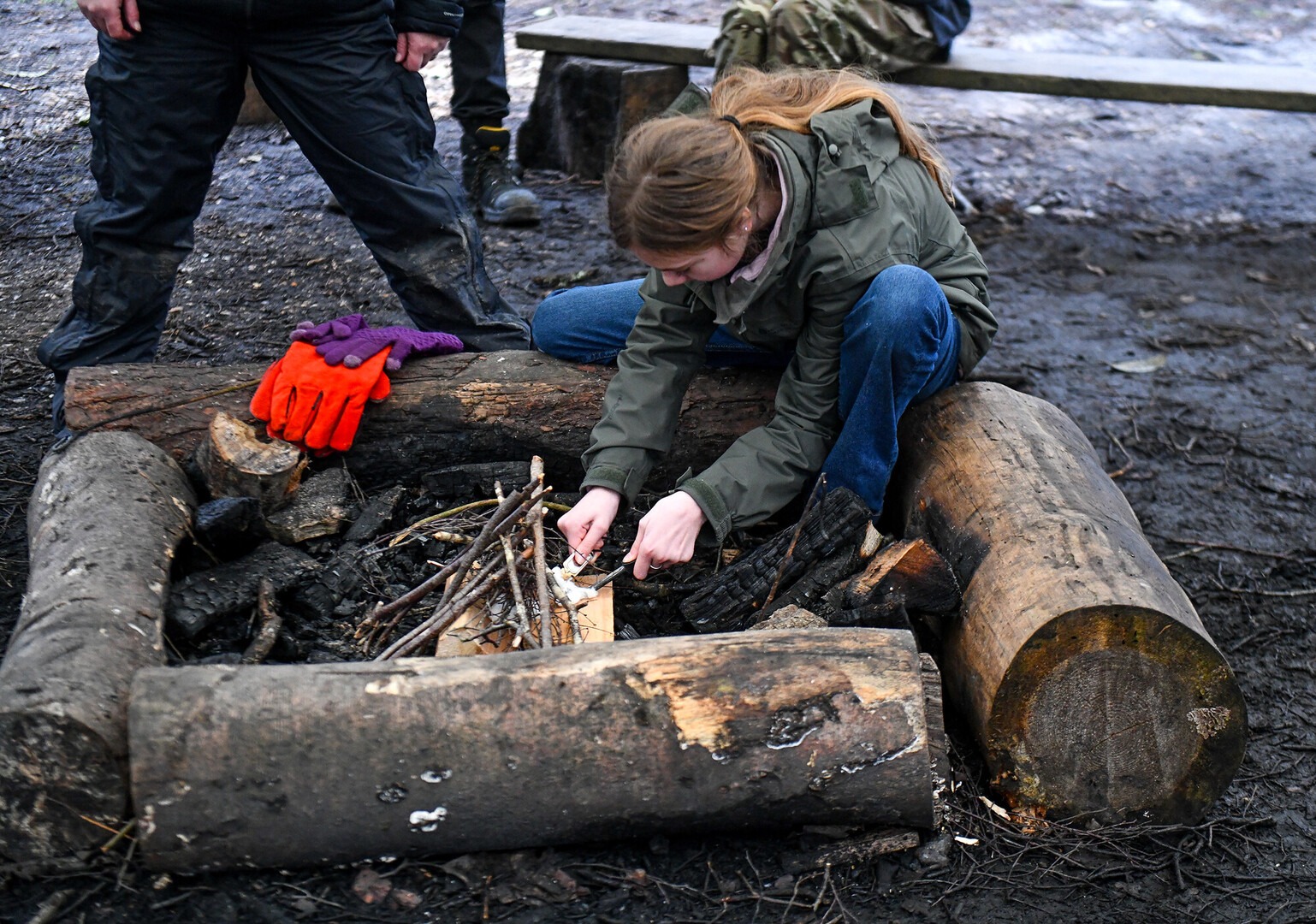 a teen sitting on a log lighting a fire in the outdoors