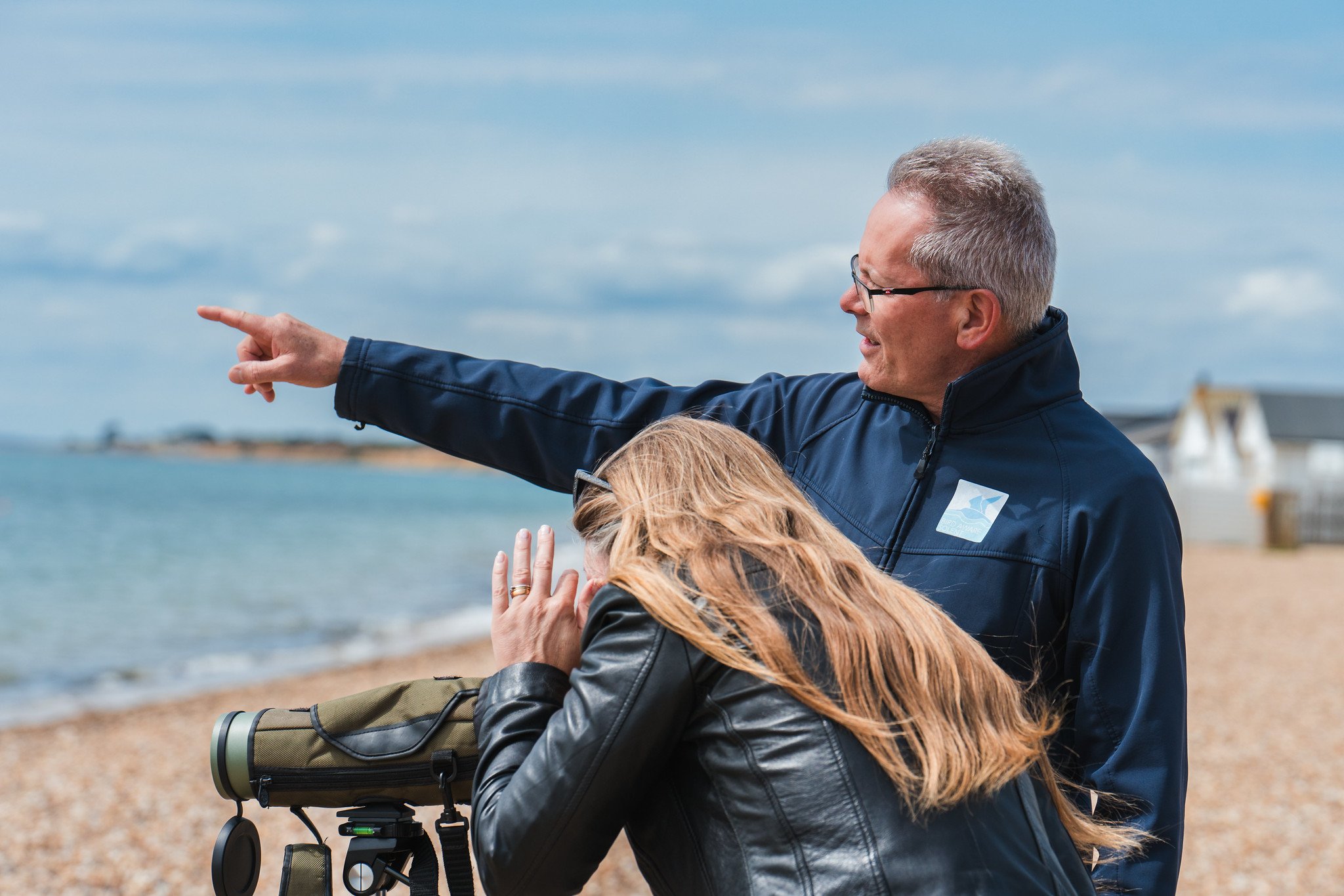A woman looking out to sea through a spotting scope next to a Bird Aware Solent staff member who is pointing at something