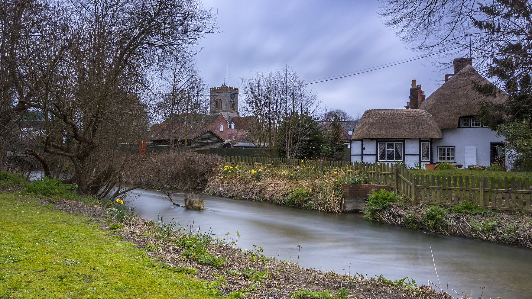 A river runs past a New Forest town, with a thatched cottage on its banks, and a church in the background