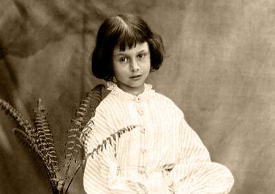 Sepia portrait of Alice Liddell seated on a striped table beside a potted fern, wearing a light dress with short bob haircut