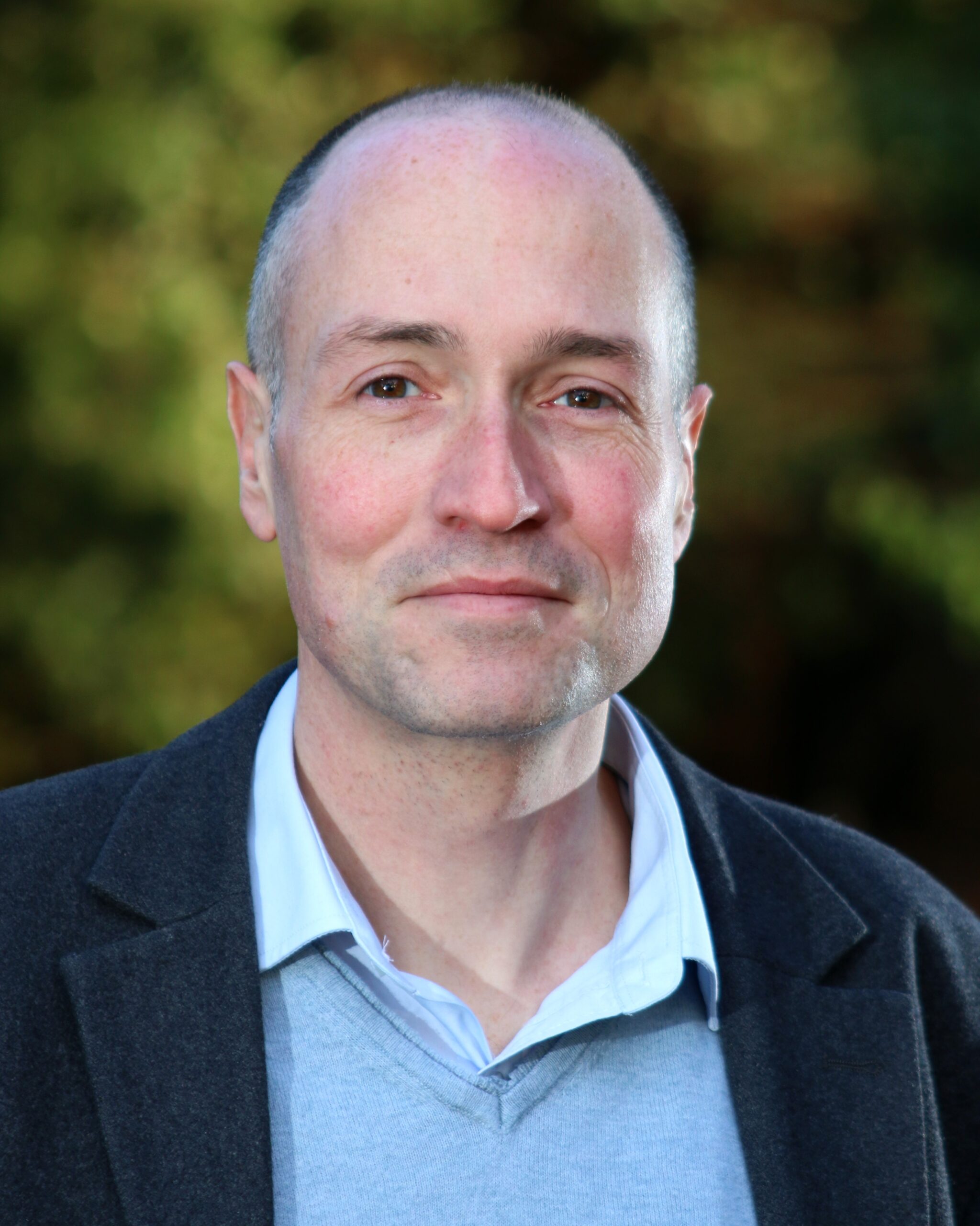 Head and shoulders of a man wearing a blue jacket and shirt with trees behind