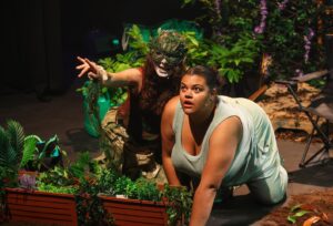 A theatre performance. A woman is bent down in front of some plants. A mystic creature appears next to her pointing at something.