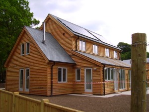 Two-story wooden house with solar panels, windows, and a fenced yard surrounded by trees
