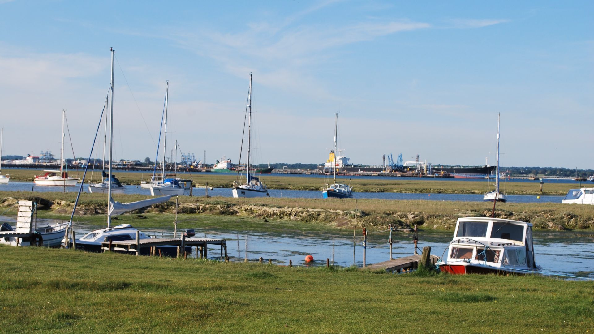Sailboats and a small motorboat moored along Ashlett Creek at low tide, with distant docks and ships under a blue sky