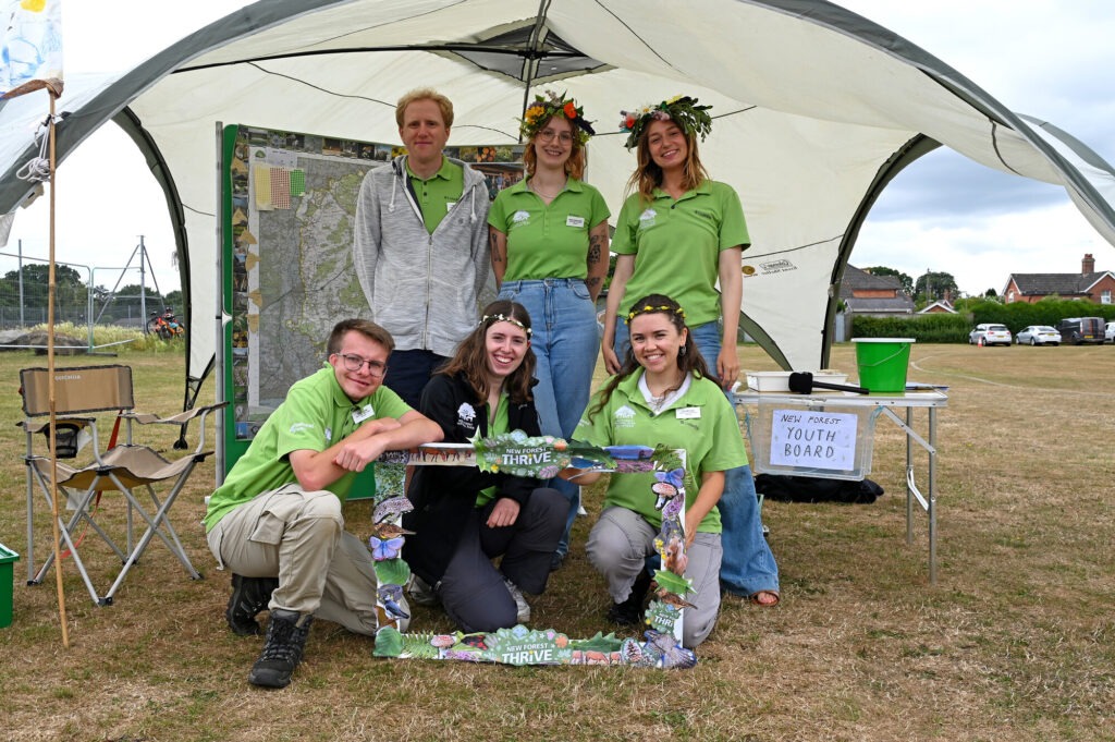 New Forest Youth Board members in green shirts at Thrive Festival booth, posing under a canopy with a New Forest Thrive frame
