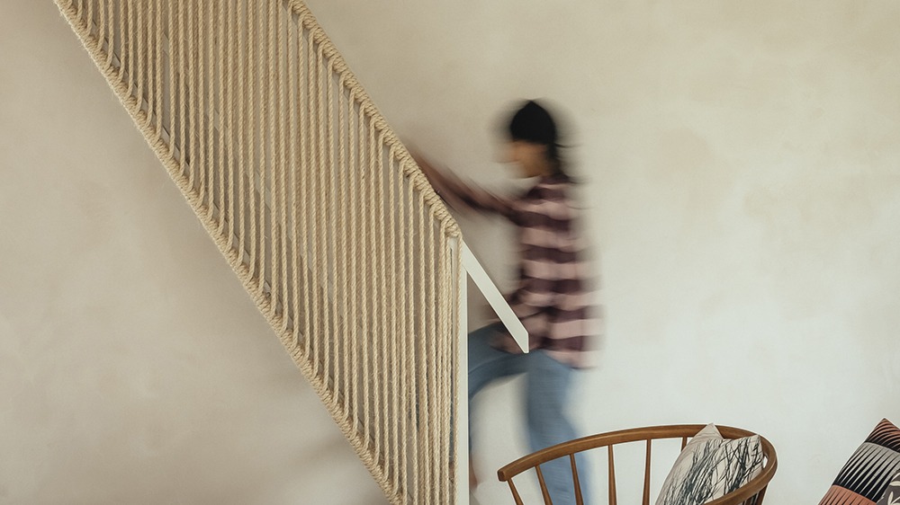 Blurred person ascending stairs with rope balustrade in a minimalist interior, with a wooden chair and cushions in foreground