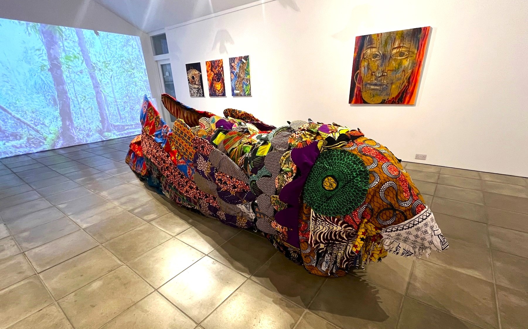 Nightjar bird sculpture covered in colorful patterned textiles, displayed in a gallery with wall artworks and forest video projection