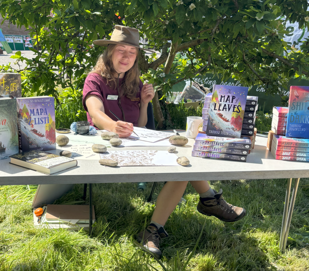 Yarrow Townsend drawing at an outdoor table under a tree, with stacks of their books The Map of Leaves and A Ship in the Dark