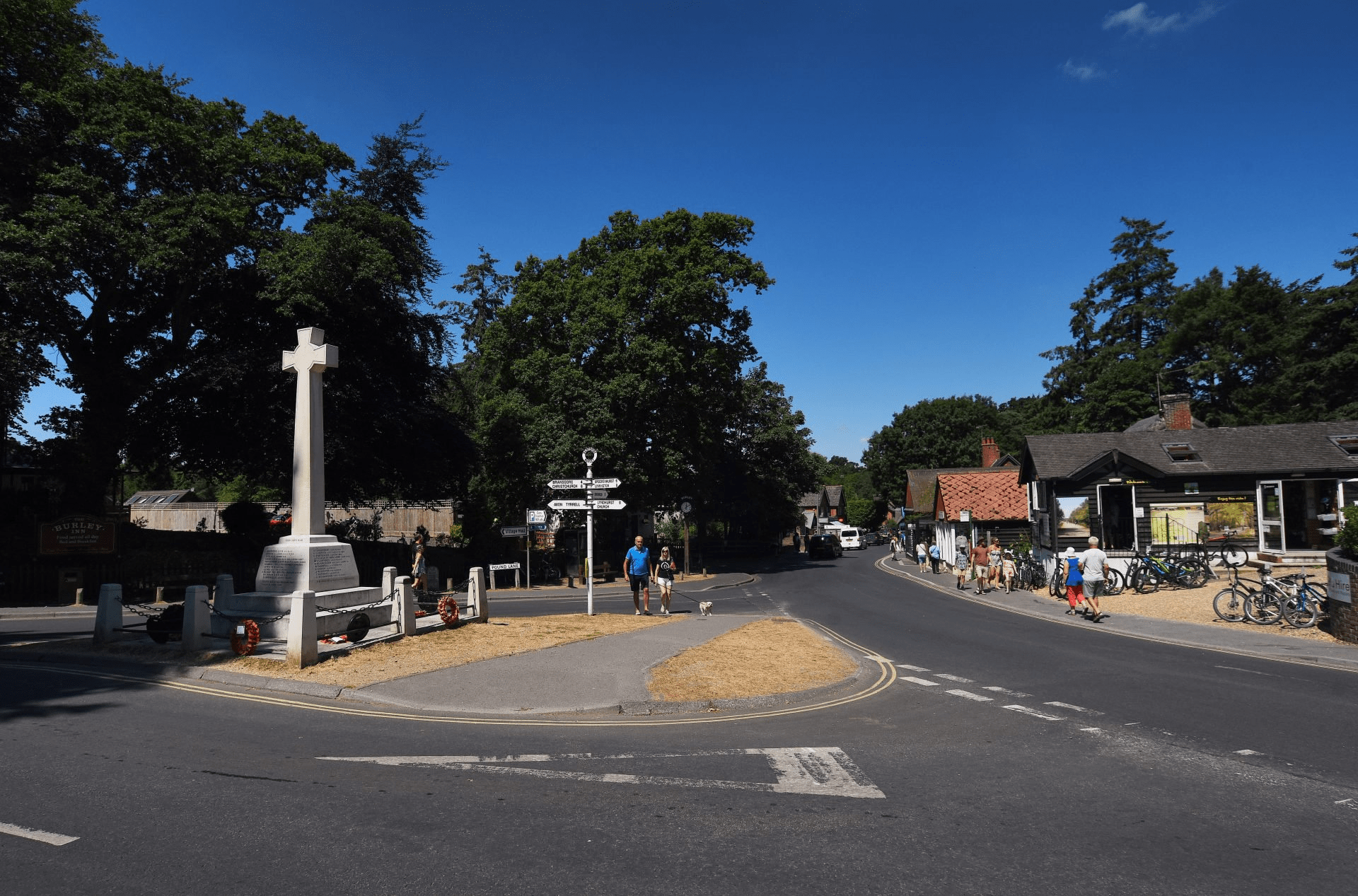 Village crossroads in Burley with a white war memorial cross, direction signpost, pedestrians, and bicycles outside shops under blue sky