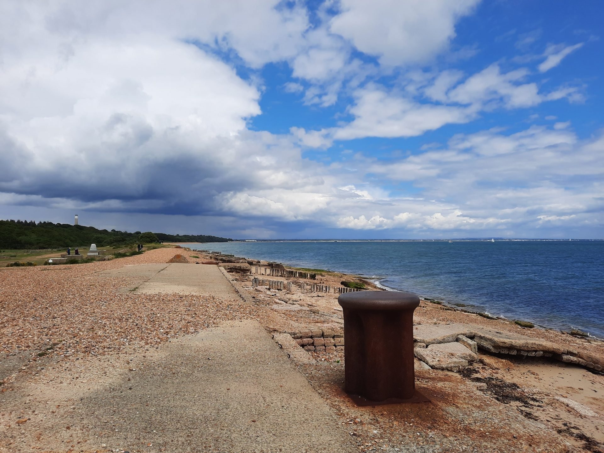 Coastal path along a shingle beach with a rusted mooring bollard in foreground, sea to the right under dramatic clouds