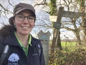 New Forest NPA west area ranger Leanne smiling beside a wooden signpost and New Forest National Park marker in woodland edge