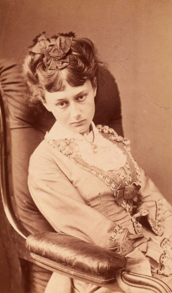 Sepia portrait of a young woman seated in an armchair, wearing a lace-trimmed dress and bow in her hair, looking at camera