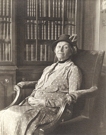 Elderly woman in a polka-dot dress and hat seated in an upholstered armchair, with bookshelves in the background