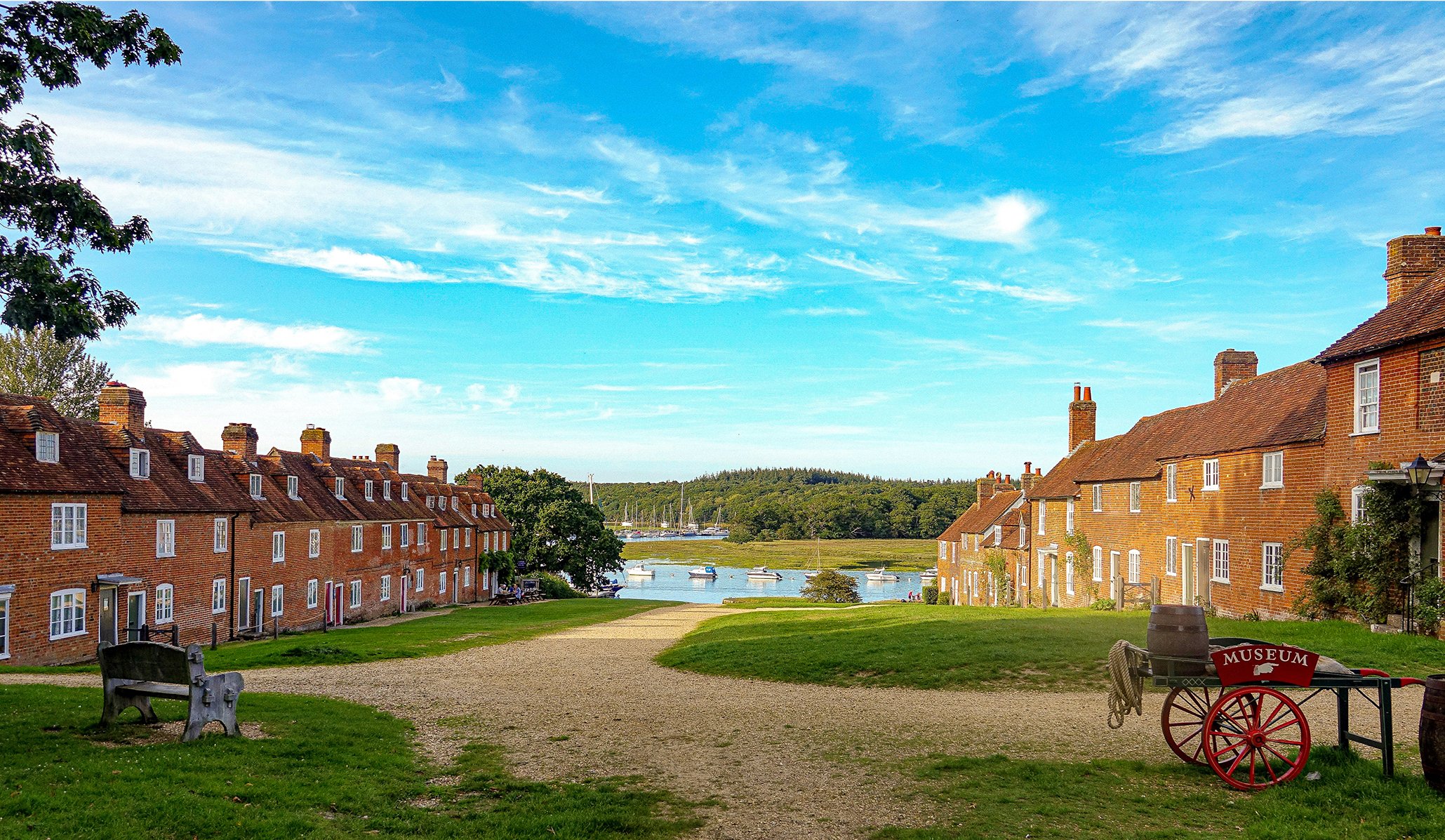 Gravel path between rows of red brick cottages leading to a river with moored boats; museum handcart in foreground