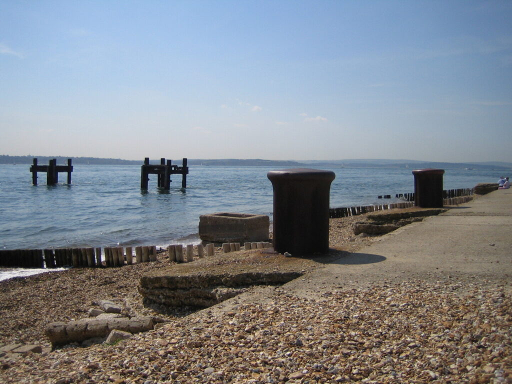 Rusty steel bollards and concrete blocks on Lepe shoreline, with old harbour supports standing in the water under a clear sky