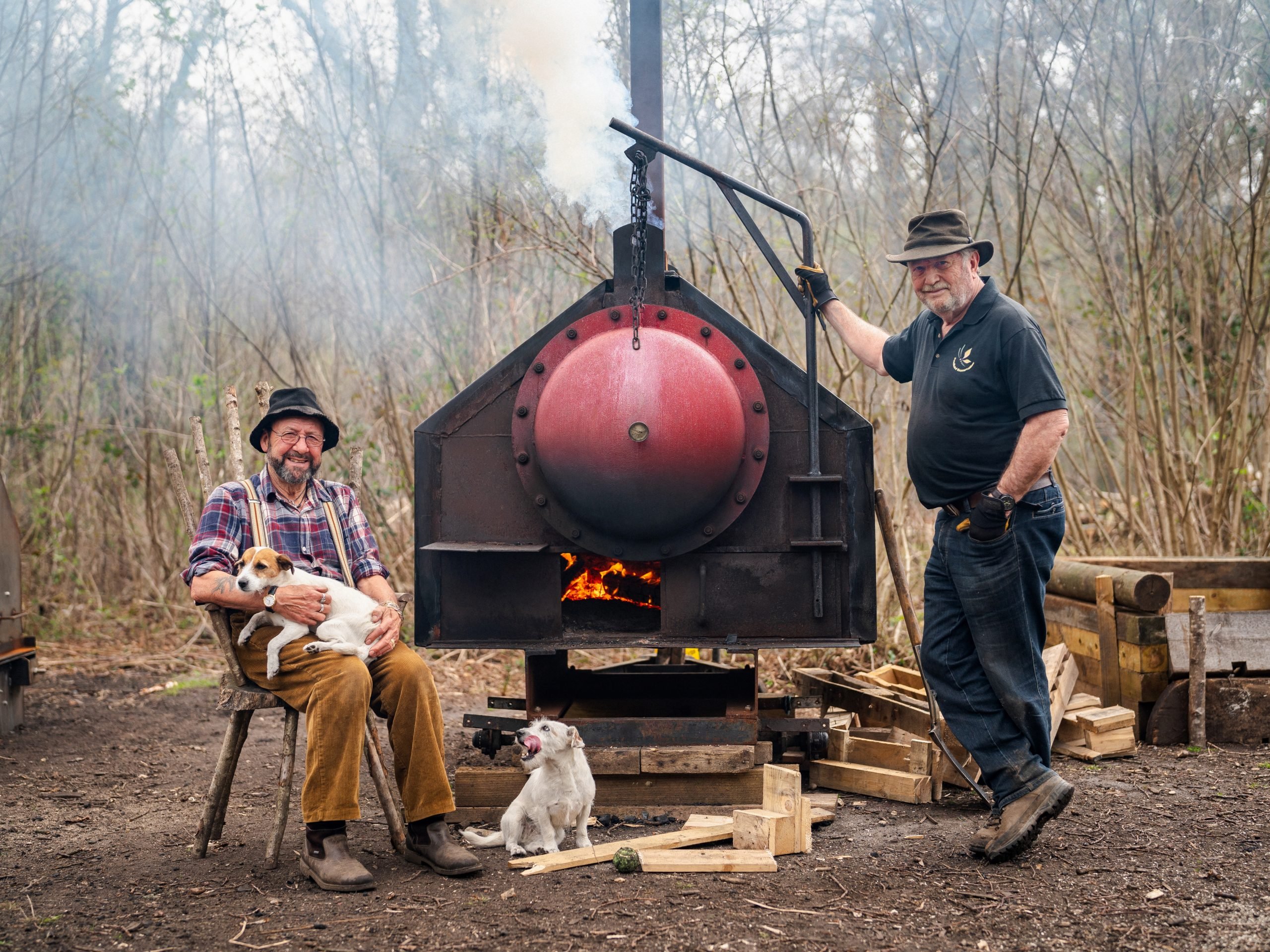Dave Dibden (left) sat with two dogs, Derek Tippetts (right) standing and leaning on the handle of a charcoal burner situated in between them but slightly behind