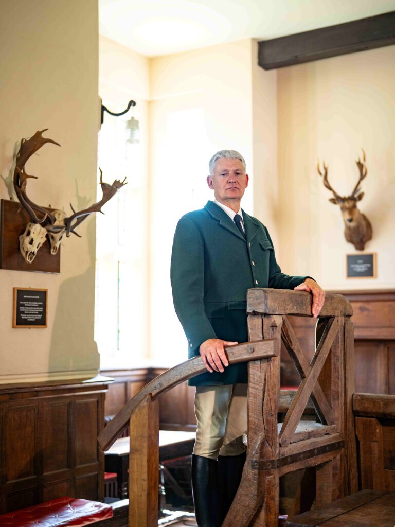 Man in green riding coat and tall black boots stands by wooden stairs in a room with mounted deer antlers