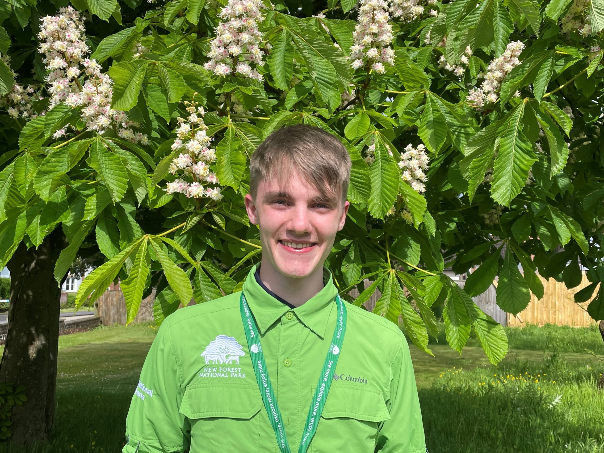 National Park Intern Josh, he is stood in front of a tree, wearing a green NPA shirt and smiling.