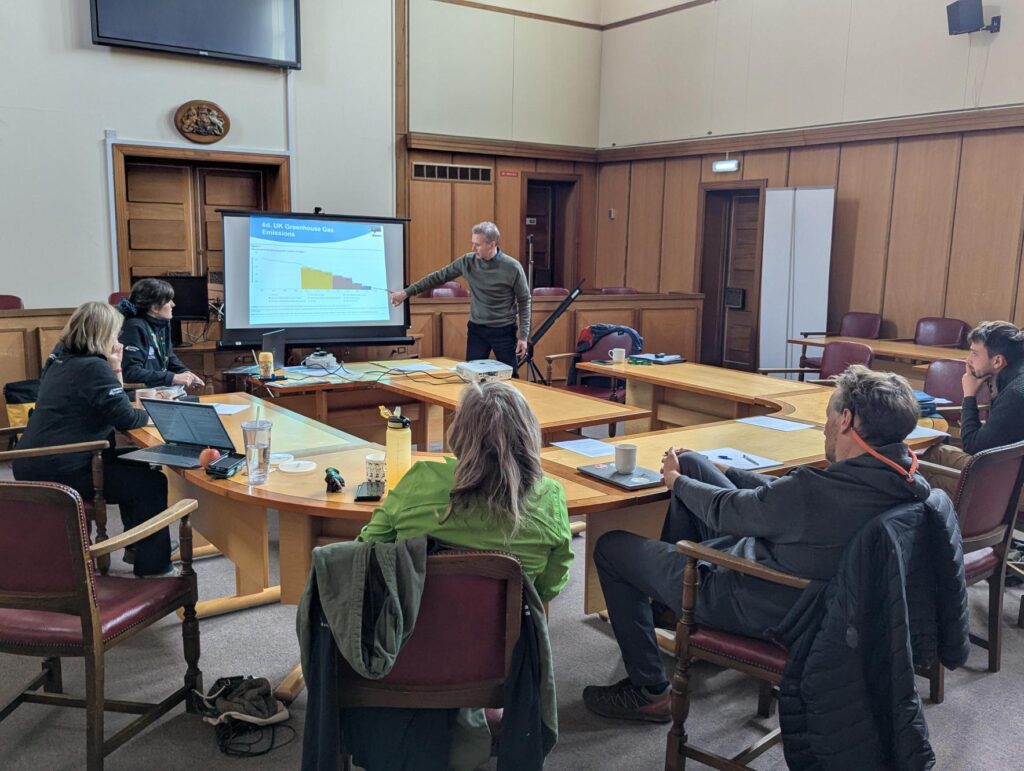 A man pointing and talking about a slideshow in front of several others sat at desks