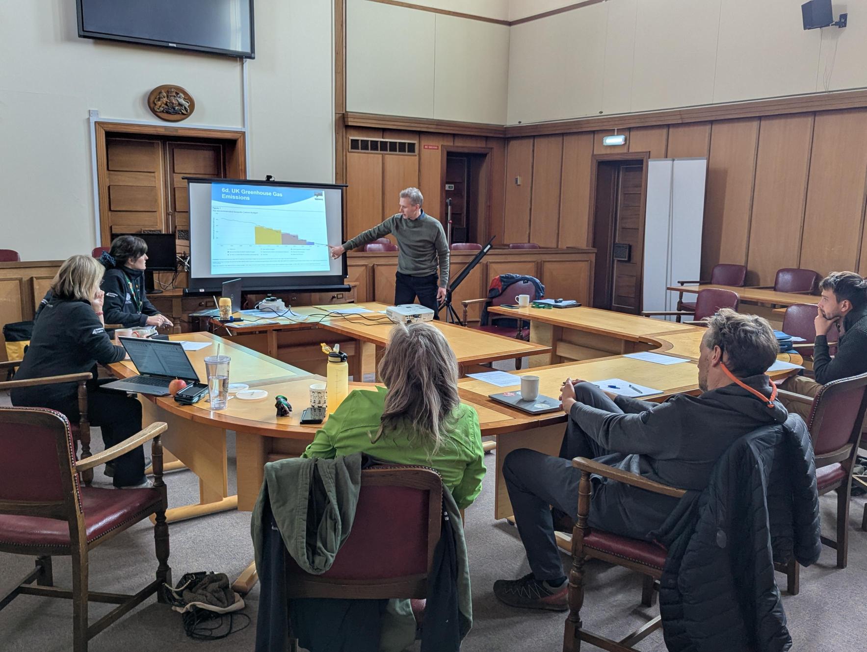 A man pointing and talking about a slideshow in front of several others sat at desks