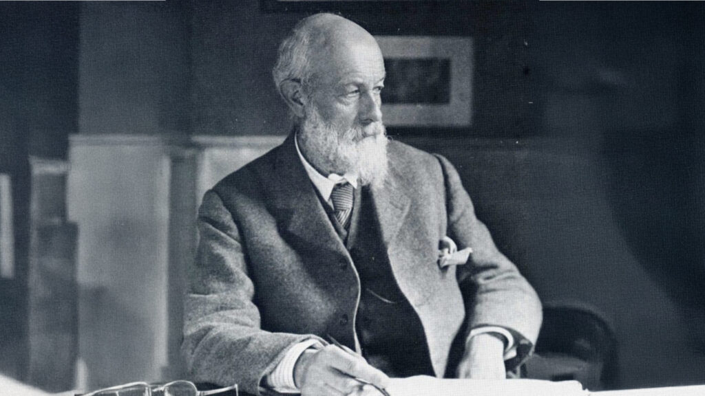 Black-and-white portrait of an older bearded man in a suit, seated at a desk holding a pen and looking to the side