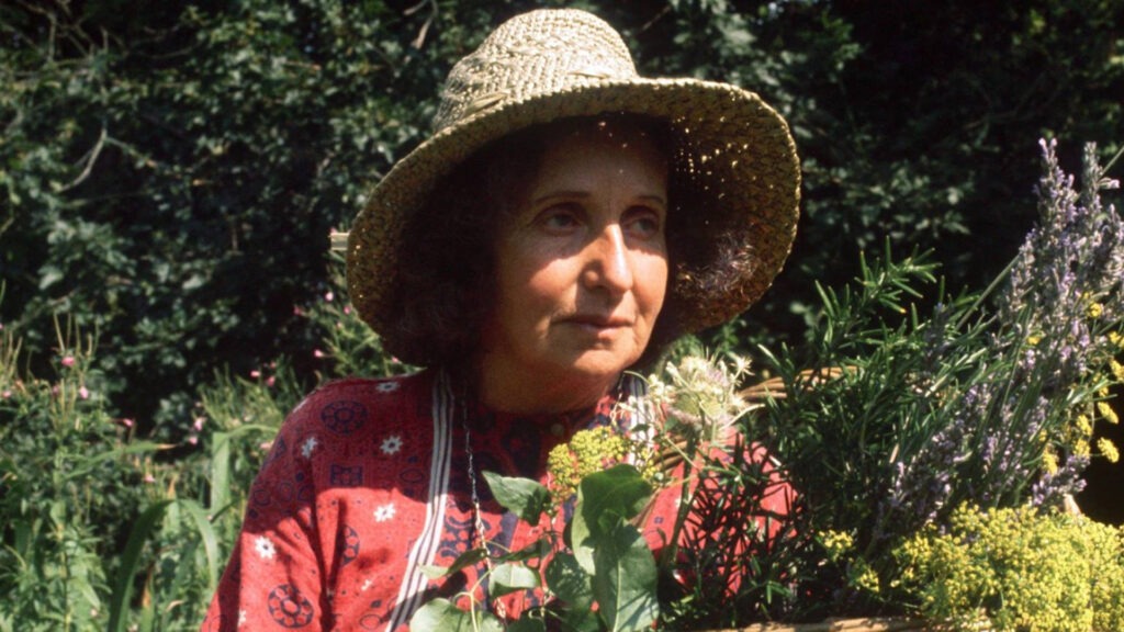 Woman in a straw hat and red patterned blouse holding a basket of herbs and wildflowers in a garden