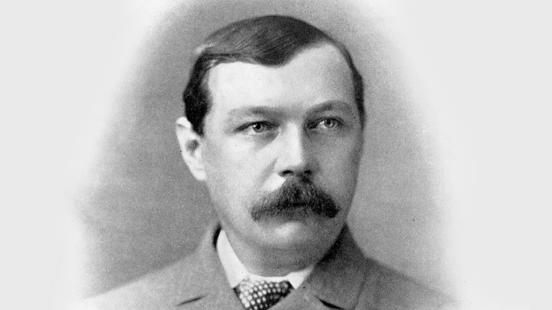 Black-and-white portrait of a mustached man in a suit and tie, facing slightly right against a plain background