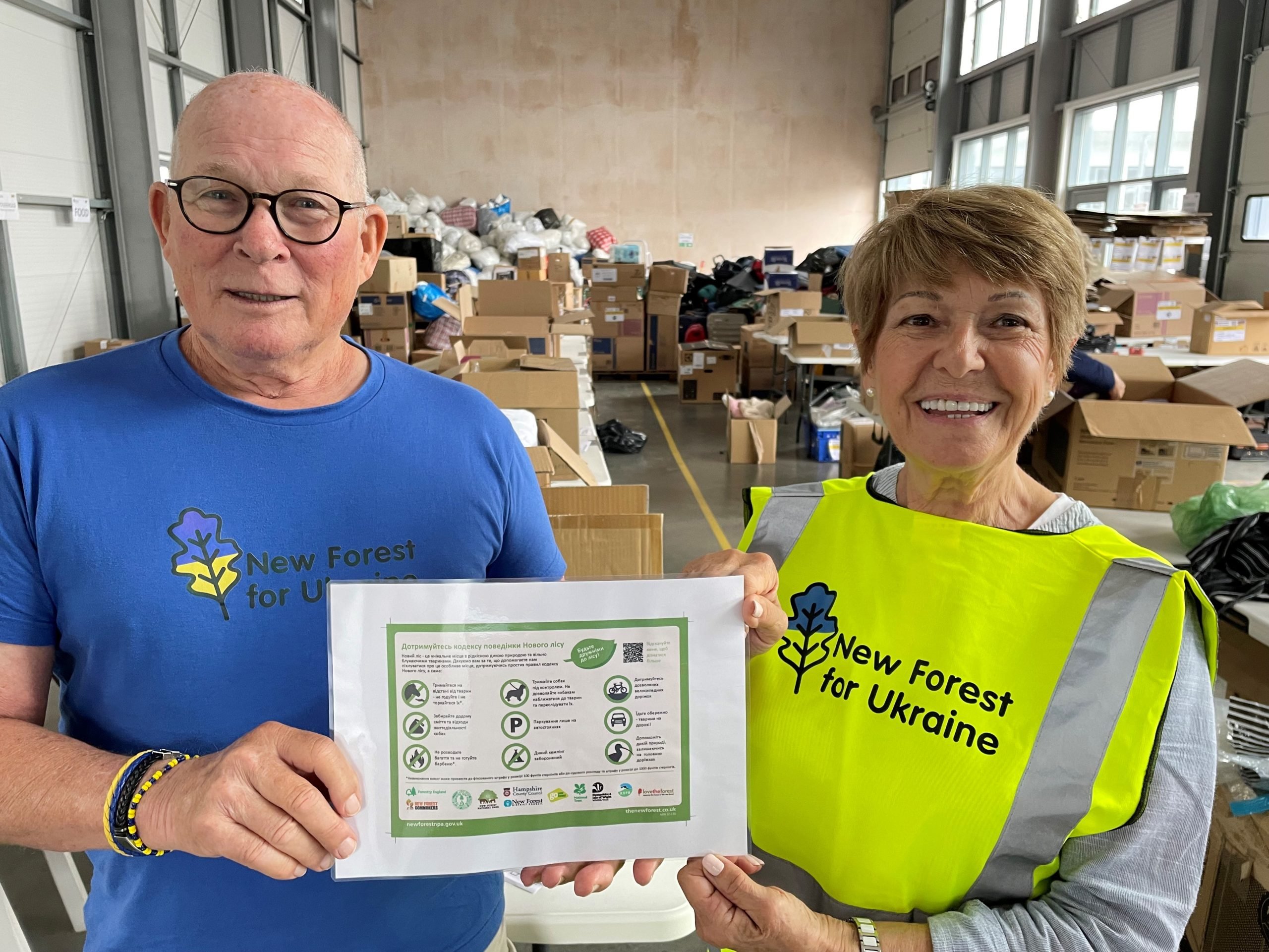 Two New Forest For Ukraine volunteers holding a Ukrainian version of the New Forest code printed out. They are stood in a warehouse full of supplies collected for Ukraine. John Allison is a bald man with glasses wearing a blue t-shirt with the New Forest For Ukraine logo on the front. Fabi is a woman with brown hair wearing a high-vis, florescent yellow bib with the New Forest For Ukraine logo on the front. Both are smiling.