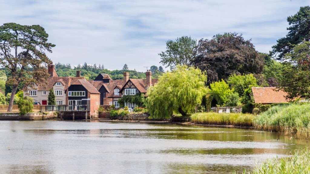 Riverside village houses with red-tiled roofs and chimneys, surrounded by trees and reeds along a calm river under cloudy sky