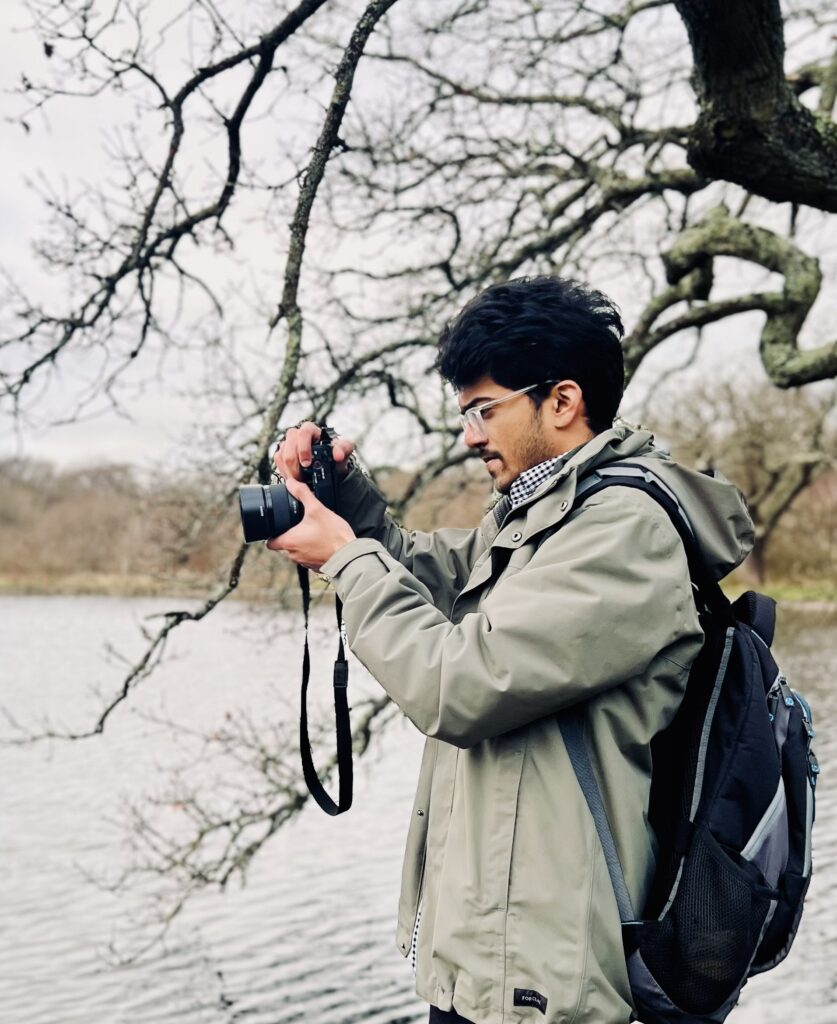 New Forest Young Content Creator Omkar Dherange photographing a lake, wearing glasses, a green jacket and a backpack under bare tree branches