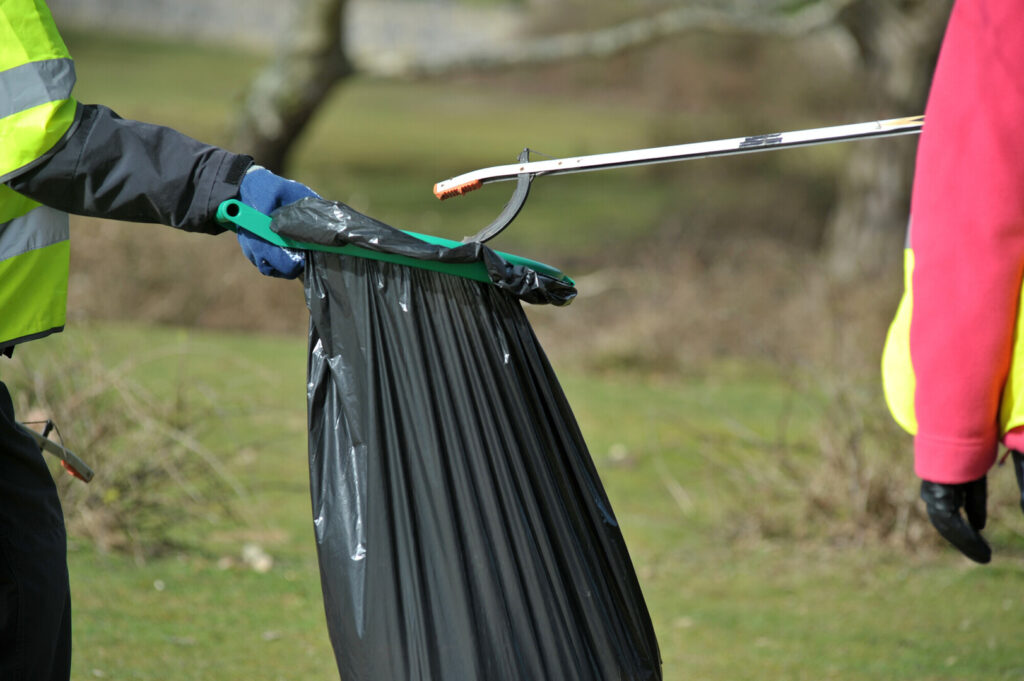 A person holds out a black bag while another person with a litter pickers puts rubbish in, set against a background of fields.