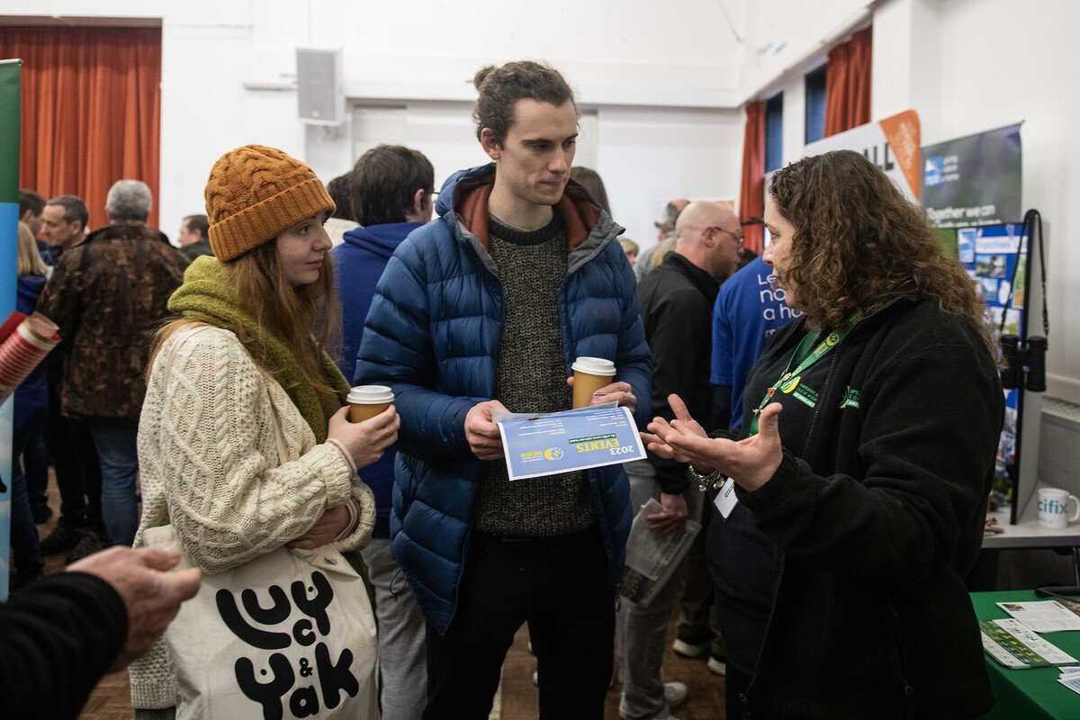 A man with a bun and a woman with a hat talking to a stall holder at the New Forest volunteer fair 2023. Both are holding coffee cups, the man also has a leaflet from one of the stalls