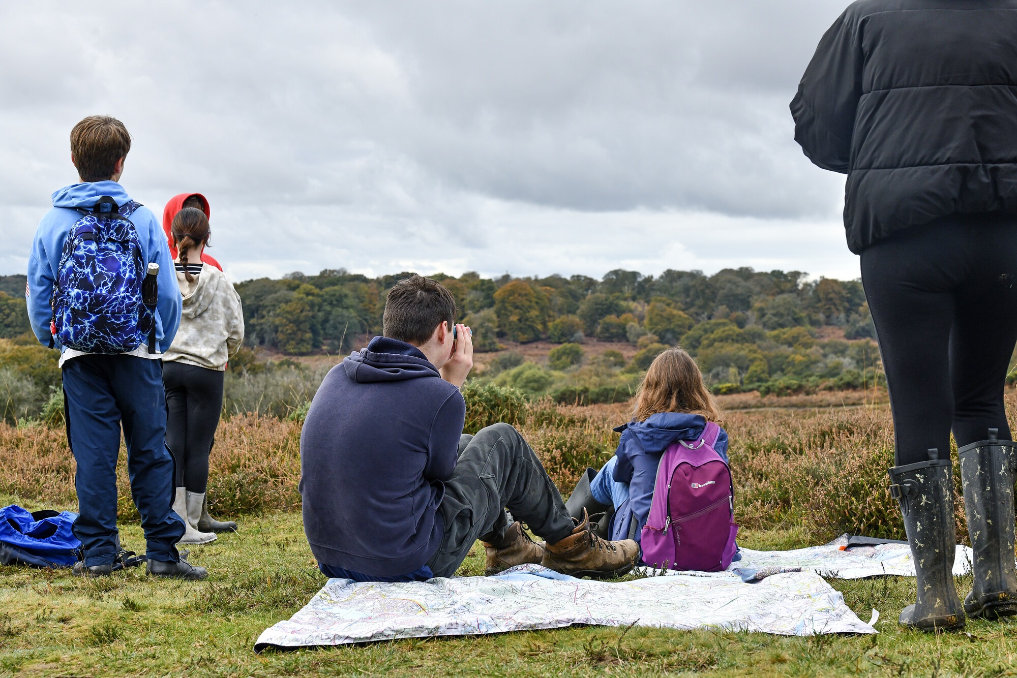 Some young people sat on a picnic blanket and others standing looking out at a viewpoint in the New Forest