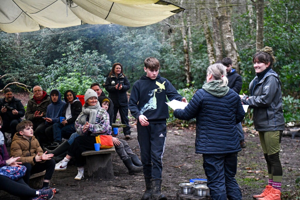 Boy receives a certificate at a Junior Rangers awards session as seated participants applaud in a wooded outdoor area