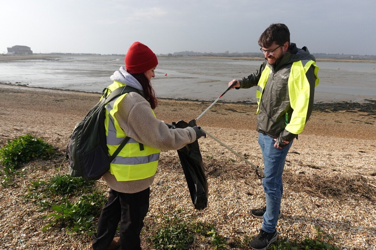 A man in high vis with a litter picker placing a piece of rubbish in a bin bag held by a woman in high vis. They are on a shingle beach on an overcast day. The woman is wearing a woolly hat.