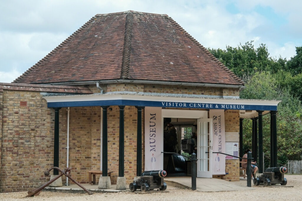 Brick visitor centre and museum with open doorway, cannons and anchor outside, and a person seated near the entrance