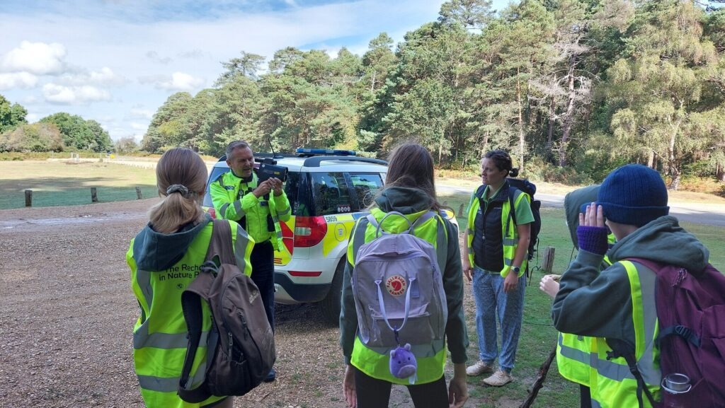 Junior Rangers in hi-vis vests watch a Hampshire Constabulary PCSO demonstrating a speed gun by a police car during an outdoor safety session